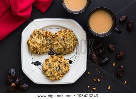 Oats and date fruit cookies in elegant white plate with two cups of tea on black table with red serviette - Top view food background