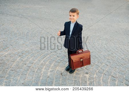 Concept future businessman. High angle of businessman in formal wear standing on pedestrian street carrying a braun briefcase looking to camera, urban gray pavage stone background. OK sign.