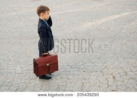 Concept future businessman. Side angle of  smiling businessman in formal wear standing on pedestrian street carrying a braun briefcase talking on the phone, urban gray pavage stone background.