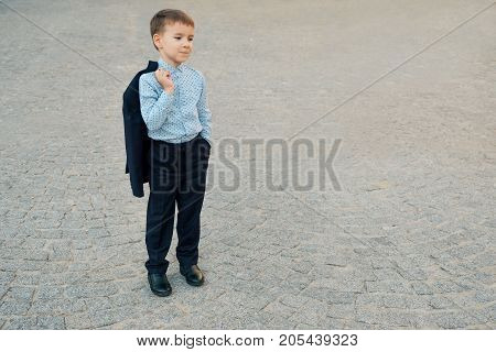 Portrait elegant little man in fashionable navy blue shirt holding dark blue jacket on his back smiling and looking to camera on urban gray pavage. Young boy posing. Boy ready for school.