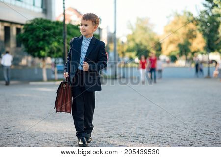 Concept future businessman. Business meeting. Low angle of future businessman in formal wear walking around the street carrying a braun briefcase and holding jacket with arm, urban background.