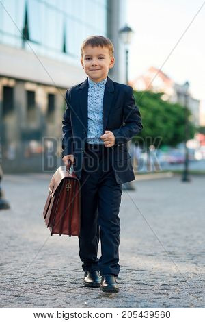 Concept future businessman. Business meeting. Portrait of future smiling businessman in formal wear walking around the street carrying a braun briefcase and holding jacket with arm, urban background.