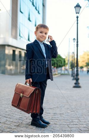 Concept future businessman. High angle of  smiling businessman in formal wear standing on pedestrian street carrying a braun briefcase talking on the phone, urban gray pavage stone background.