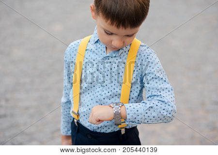 Concept future businessman. Boy portrait in dark blue pants, baby blue shirt and yellow straps, standing on pedestrian street with braun briefcase looking to his watch, urban background.