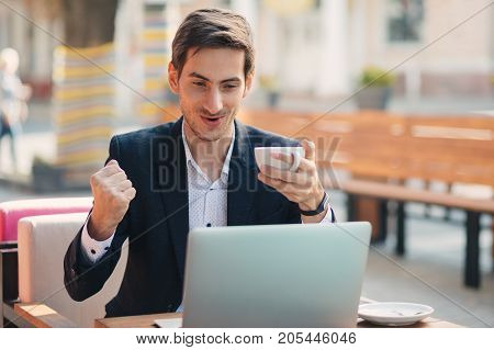 Emotions concept. Gyu with winner look sitting at table holding cup of coffee in hand looking to laptop. Young man is happy to have the contract signed, to get the email with job acceptance.
