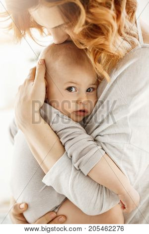 Portrait of sweet little child looking in the camera with interested expression while mother tenderly snuggle palming and kissing her newborn baby