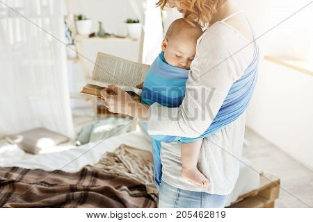 Tiny sweet babyboy peacefully sleeping on mother in baby sling while young beautiful mom trying to relax reading favorite book