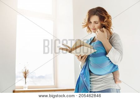 Happy beautiful mother snuggles her napping child to chest and reading book about child education in bright cozy room in front of window. Family moments