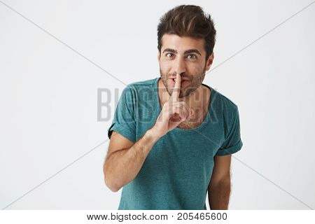 Studio portrait of funny good looking caucasian male in blue t-shirt. with good hair and beard, smile while telling secret to his friend and asking not to tell anybody