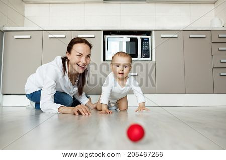 Joyful beautiful mother and little baby son with big brown eyes playing together with red ball in kitchen. Scene of family happiness