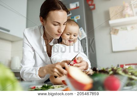 Serious young good-looking mother spending time with her newborn son cooking dinner and showing how to cut vegetables