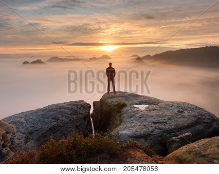 Man stands alone on the peak of rock. Hiker watching to autumn Sun at horizon . Beautiful moment the miracle of nature. Colorful mist in valley. Man hike. Person silhouette stand