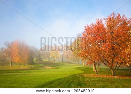 Morning mist and sun light. Fog over golf course during beautiful fall sunrise. Colorful autumn trees on a bright green lawn. Horizontal composition.