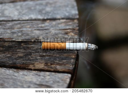Burning cigarette with ash and smoke left on the outdoor wooden table. Bad habit concept background.