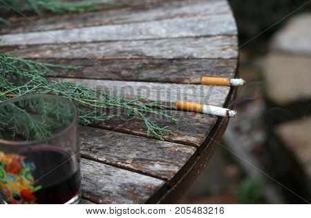Burning cigarette with ash and smoke in focus left on the outdoor wooden table next to almost empty glass with wine and another cigarette stub out of focus. Bad habits concept background.
