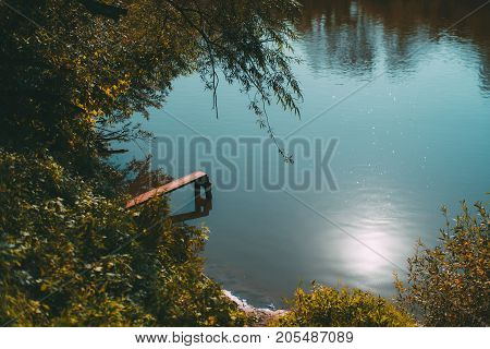 Autumn pond reflecting teal sky with small wooden springboard for jumping into the water; greenery around bushes sludgy riverside trailing tree branches sunny day Russia