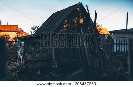 Dark silhouette of abandoned wooden house in holiday village on sunset with strong sun spots on the roof; blue clear sky fencing and other houses in background; decomposed beams leant on facade