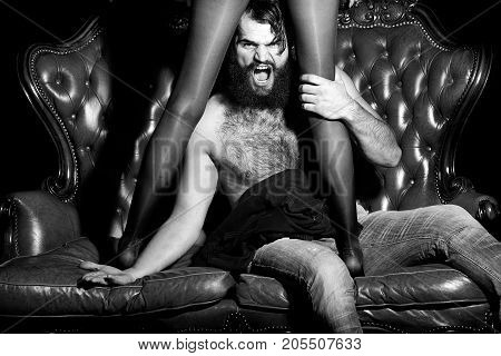 One handsome serious young adult man with long black lush beautiful beard moustache and bare hairy chest sitting on leather couch with sexy female legs in stockings indoor horizontal picture