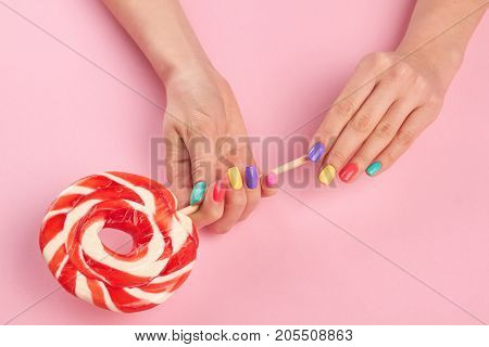 Bright lollipop in female hands. Female hands with multicolor summer nails holding tasty big lollypop on pink background, top view.