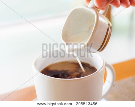 Closeup woman hand pouring hot milk into black coffee cup during breakfast time with selective focus.