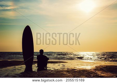 Silhouette of surf man sitting with a surfboard on the seashore beach at sunset time. Handsome Asia man model in his 20s.