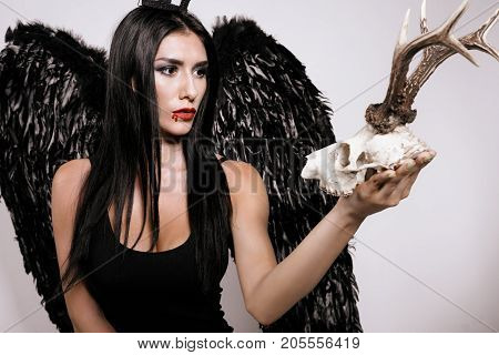 The demon girl. The black-haired model in a black dress and with black wings behind her pensively looks at the deer skull she holds in her hands
