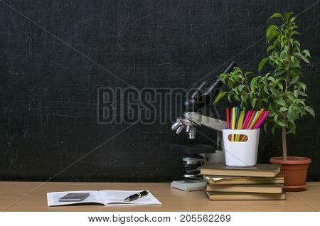 Teacher or student desk table. Education background. Education mockup concept. Microscope open copybook stacked books plant in the pot and colour pencils on blackboard (chalkboard) background.