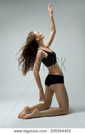 Slim brunette her hair loose wearing black sporty top and tanga staying on her knees rearview on gray background