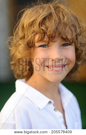 Portrait of the nice boy of 10-11 years. The fellow has beautiful fair curly hair blue cheerful eyes suntanned skin a pleasant smile. The boy is dressed in a white shirt.