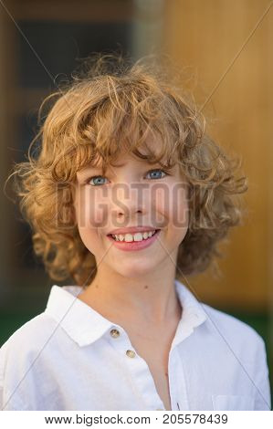 Portrait of the nice boy of 10-11 years. The fellow has beautiful fair curly hair blue cheerful eyes suntanned skin a pleasant smile. The boy is dressed in a white shirt.