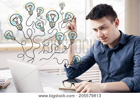 Handsome young businessman at modern workplace using laptop with aabstract drawn light bulb balloons. Achievement concept