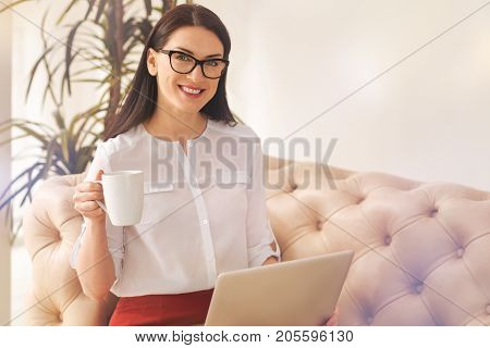Look at me. Pretty businesswoman holding laptop on knees and keeping smile on face while posing on camera