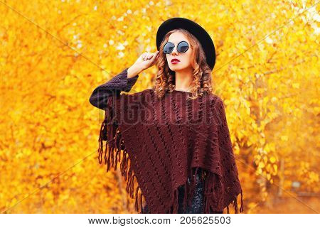 Autumn Fashion Woman Wearing A Black Round Hat, Knitted Poncho On A Yellow Leaves Background