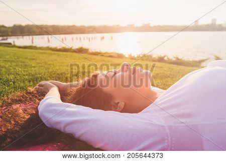 Beautiful young pregnant woman doing yoga on river bank. Woman lying on her back on a yoga mat. Female face closed eyes close-up. River setting sun in the background