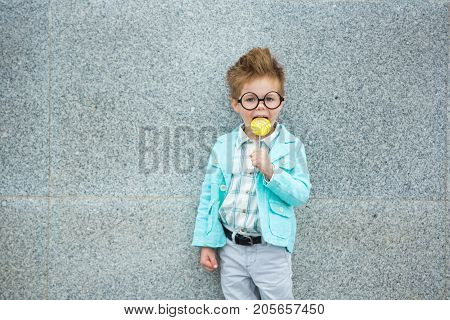 Fashion baby boy in mint jacket stands on a gray wall background. Trendy boy with lollipop standing on the street.