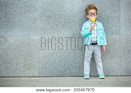 Fashion baby boy in mint jacket and sneakers stands on a gray wall background. Trendy boy with lollipop standing on the street.