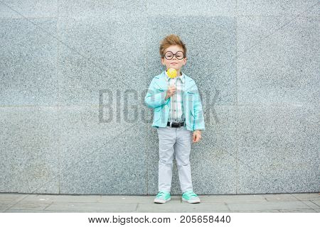 Fashion baby boy in mint jacket and sneakers stands on a gray wall background. Trendy boy with lollipop standing on the street.