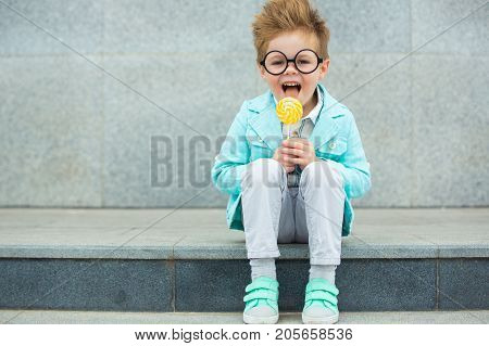 Fashion baby boy in mint jacket and sneakers stands on a gray wall background. Trendy boy with lollipop standing on the street.