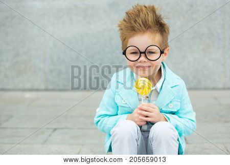 Fashion baby boy in mint jacket stands on a gray wall background. Trendy boy with lollipop standing on the street.