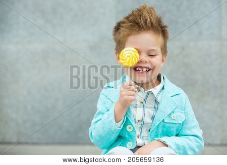 Fashion baby boy in mint jacket and sneakers stands on a gray wall background. Trendy boy with lollipop standing on the street.