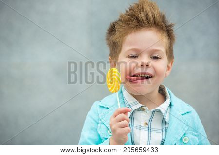 Fashion baby boy in mint jacket and sneakers stands on a gray wall background. Trendy boy with lollipop standing on the street.