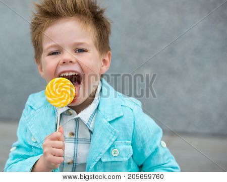 Fashion baby boy in mint jacket and sneakers stands on a gray wall background. Trendy boy with lollipop standing on the street.