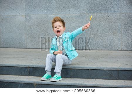Fashion baby boy in mint jacket and sneakers stands on a gray wall background. Trendy boy with lollipop standing on the street.