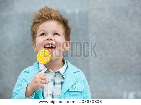 Fashion baby boy in mint jacket and sneakers stands on a gray wall background. Trendy boy with lollipop standing on the street.
