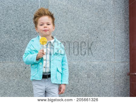 Fashion baby boy in mint jacket and sneakers stands on a gray wall background. Trendy boy with lollipop standing on the street.