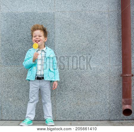 Fashion baby boy in mint jacket and sneakers stands on a gray wall background. Trendy boy with lollipop standing on the street.