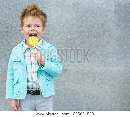 Fashion baby boy in mint jacket and sneakers stands on a gray wall background. Trendy boy with lollipop standing on the street.