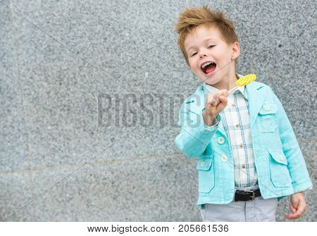 Fashion baby boy in mint jacket and sneakers stands on a gray wall background. Trendy boy with lollipop standing on the street.