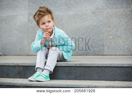 Fashion baby boy in mint jacket and sneakers posing on a gray wall background. Trendy boy standing on the street.
