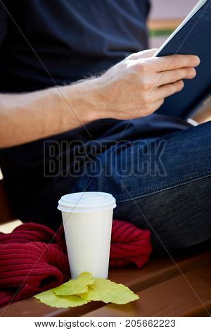 Sitting man reading a book and drinking warm tea. Cup of coffee on the bench. Close-up.